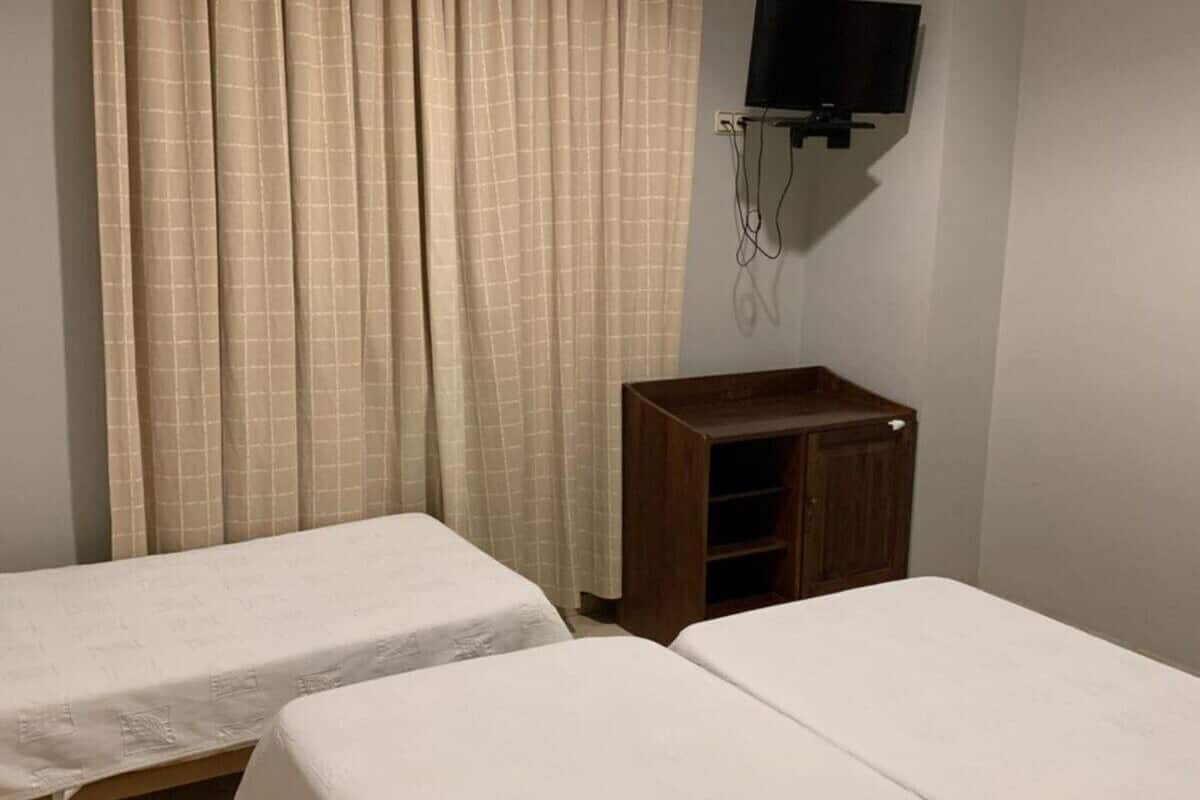 A room with two single beds, a wooden cabinet, a flat-screen TV mounted on the wall, and a window with beige curtains.
