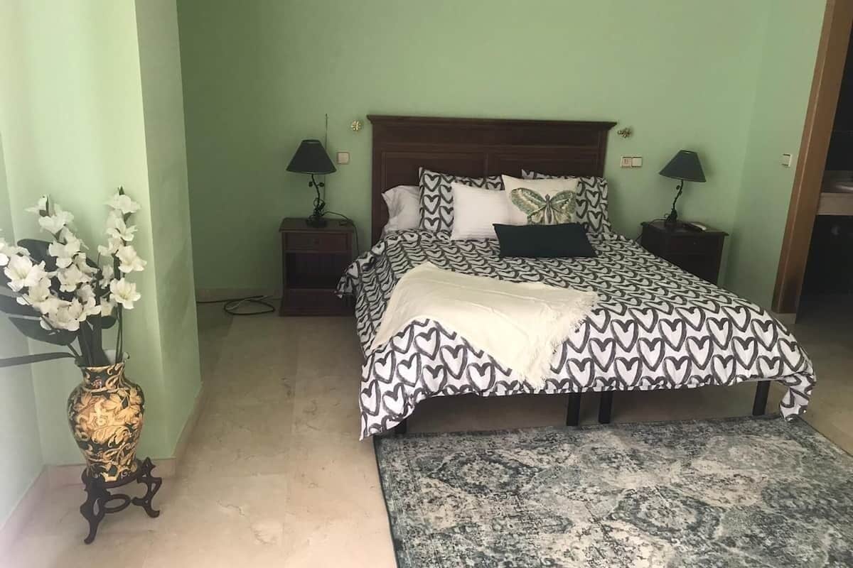 A bedroom with a bed, two bedside tables, a nightstand, a patterned rug, and a vase with flowers.