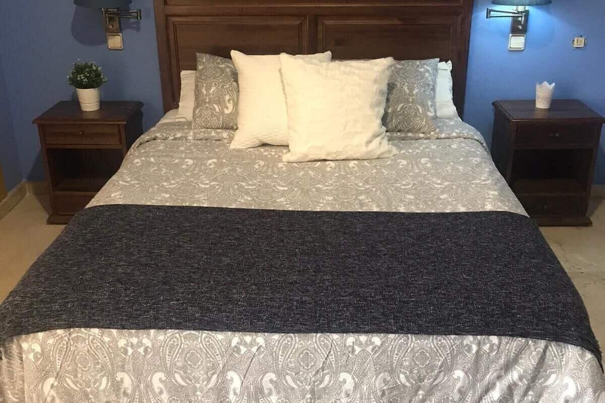 A neatly made bed with patterned bedding and a dark throw blanket. There are two bedside tables, each with a lamp and a small plant.