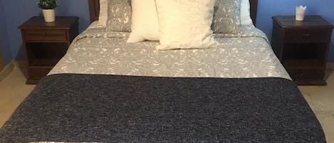 A neatly made bed with patterned bedding and a dark throw blanket. There are two bedside tables, each with a lamp and a small plant.
