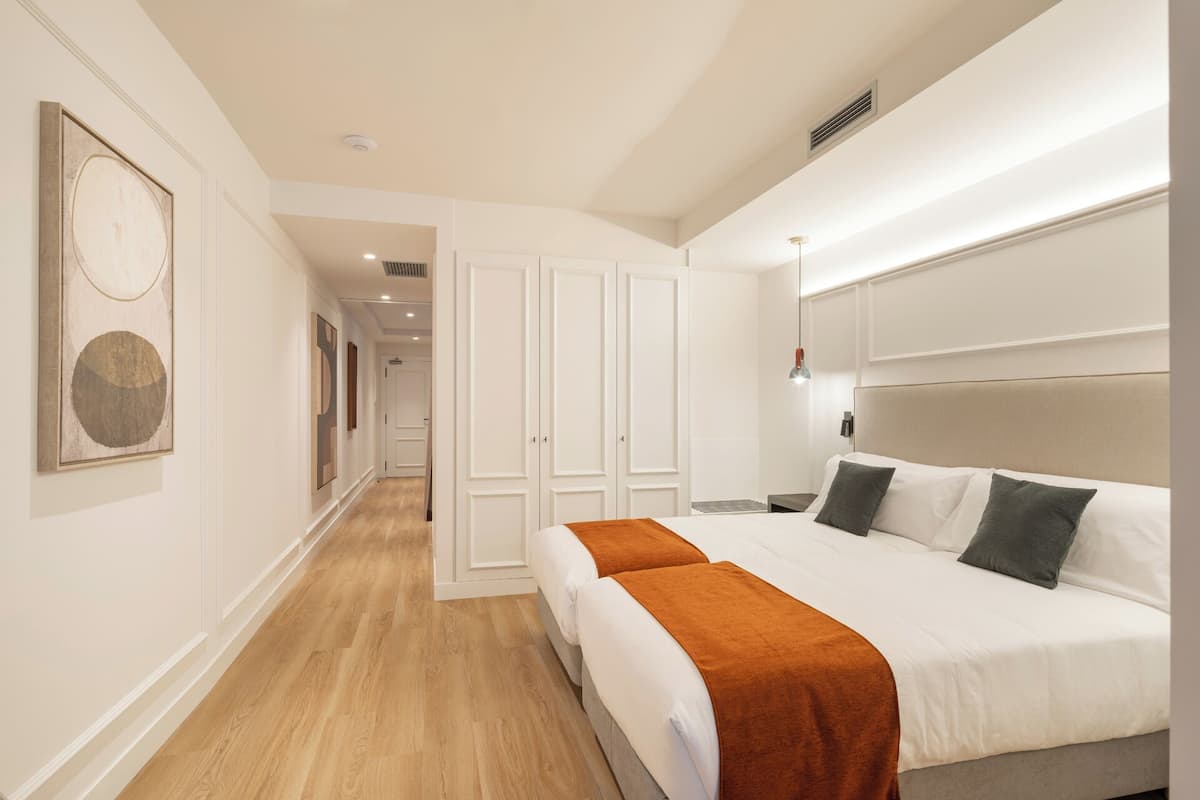 A hotel room with a large bed, a wooden floor, white walls, and a hallway leading to another room.