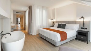 A modern hotel room with a large bed, bedside tables, a bathroom with a bathtub, and a hallway leading to a sitting area. - Marina 30 (A Coruña)