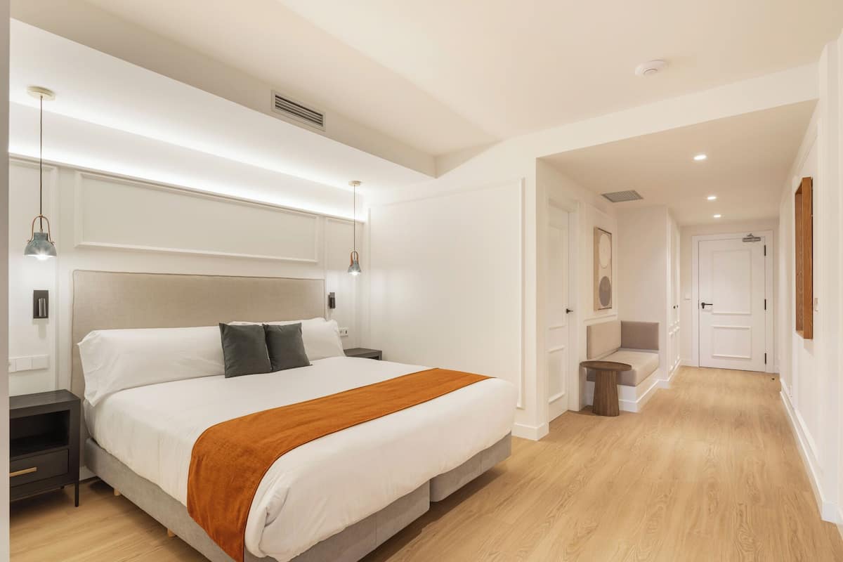 A modern bedroom with a large bed, bedside tables, a nightstand, a wooden floor, and a hallway leading to another room.