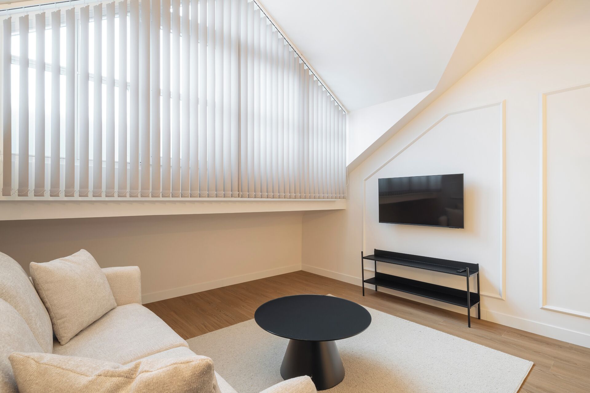 Un salon moderne avec un canapé, une table ronde noire, un téléviseur fixé au mur et une fenêtre équipée de stores.
