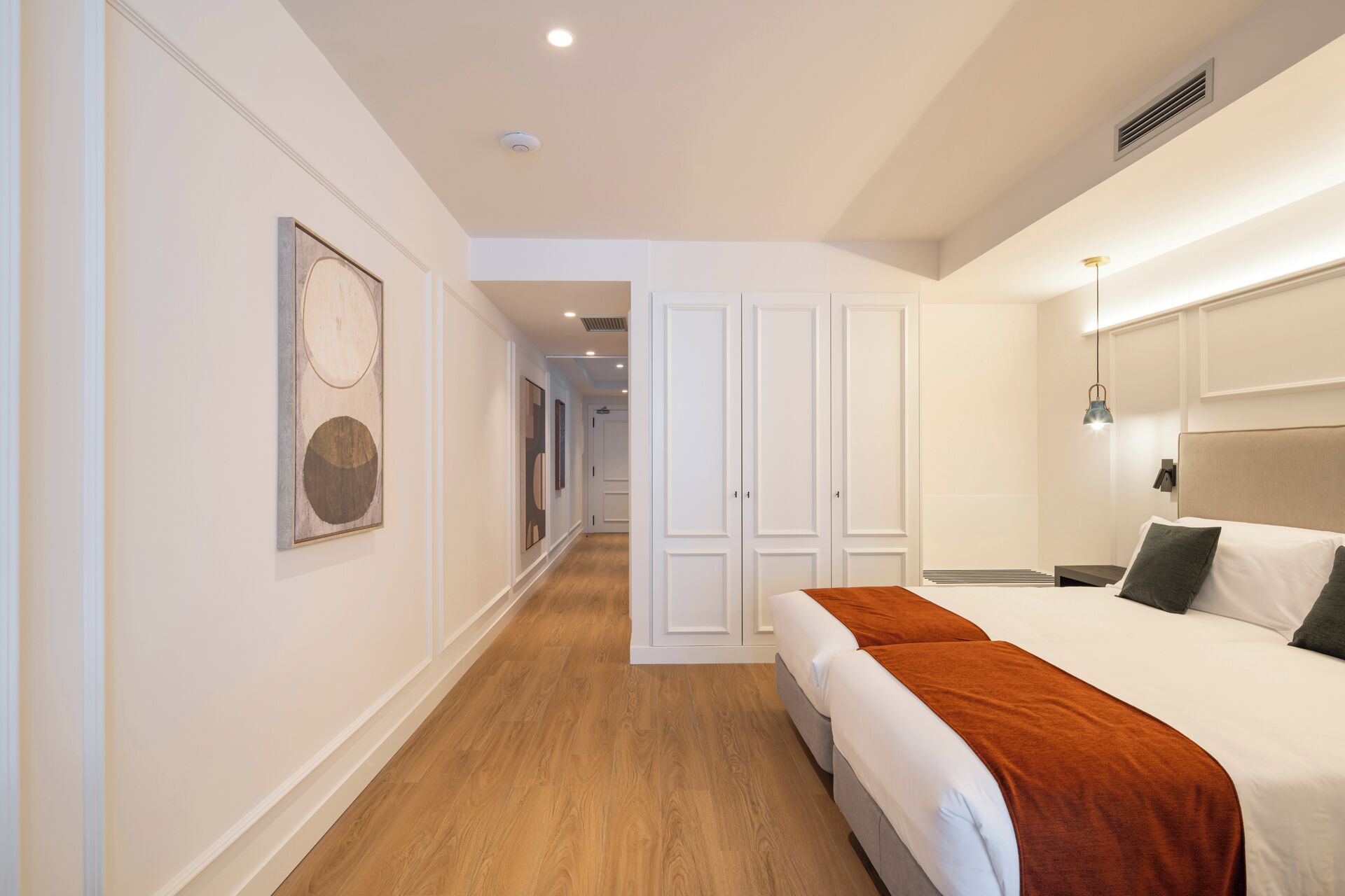 A hotel room with a bed, a nightstand, a bedside table, a wardrobe, and a hallway with a door leading to another room.