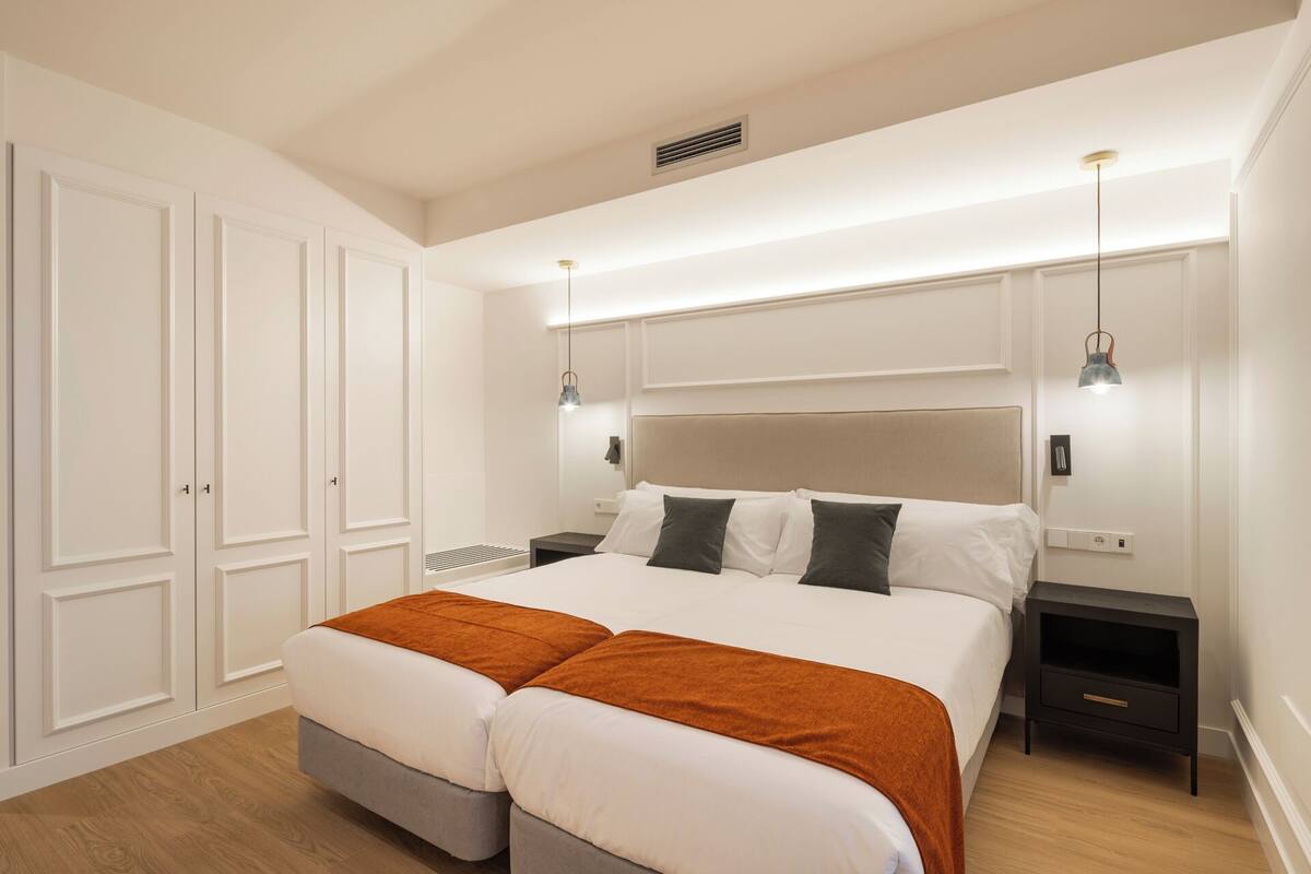 A bedroom with a double bed, two bedside tables, a wardrobe, and a ceiling vent.