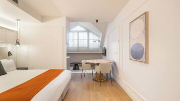 A hotel room with a bed, a small dining table, and chairs. There is a window with blinds, a painting on the wall, and a ceiling vent.