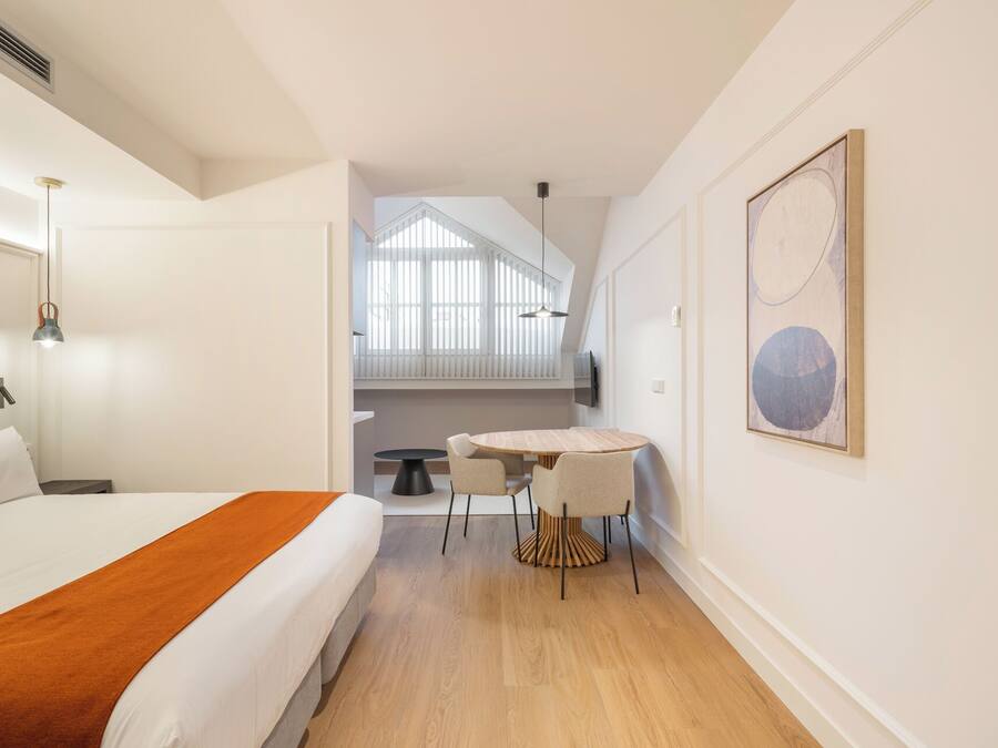 A hotel room with a bed, a small dining table, and chairs. There is a window with blinds, a painting on the wall, and a ceiling vent.