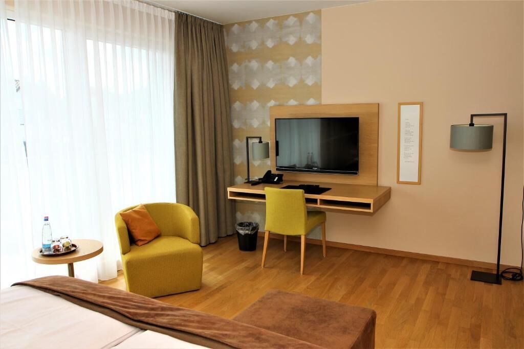 Une chambre d’hôtel avec un lit, un bureau avec une télévision, une chaise jaune, une petite table avec une bouteille et une poubelle.