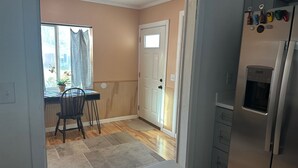 Una cocina con refrigerador, una ventana con cortinas, una mesa de comedor con sillas y una puerta que da a otra habitación.