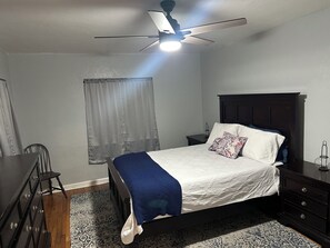 A bedroom with a bed, a nightstand, a dresser, a ceiling fan, and a window with curtains.
