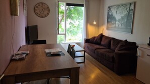 A cozy living room with a wooden table, a brown sofa, a clock, and a window with a view of greenery. - Clean bed & breakfast with roofterrace in Amsterdam! (Amsterdam)
