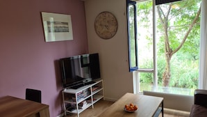 A living room with a flat-screen TV, a bookshelf, a coffee table with oranges, a clock, and a window with a view of greenery. - Clean bed & breakfast with roofterrace in Amsterdam! (Amsterdam)