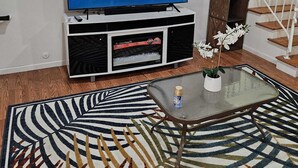 A living room with a flat-screen TV on a stand, a glass coffee table with a bottle, a patterned rug, and a staircase with white railings. - Charming 3-bedroom townhome in Pleasant Des Plaines with AC, WiFi (Des Plaines)