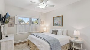 A bedroom with a bed, bedside lamps, a ceiling fan, a window with shutters, and a framed picture on the wall. - Serene Retreat on East Beach sleeps 6 guests (St Simons Island)