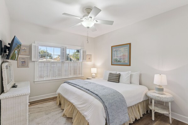 Un dormitorio con cama, lámparas de noche, ventilador de techo, ventana con contraventanas e imagen enmarcadas en la pared.
