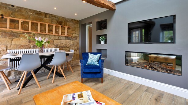 A modern living room with a stone wall, a blue armchair, a wooden coffee table, and a fireplace with a log fire. - Cherry Cottage Nr Grantley Hall (Grantley)