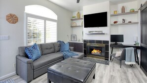 A modern living room with a gray sofa, a black ottoman, a fireplace, a flat-screen TV, and a ceiling fan.