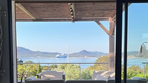A balcony with a wooden deck, two cushioned seating areas, and a view of a body of water with a boat in the distance.