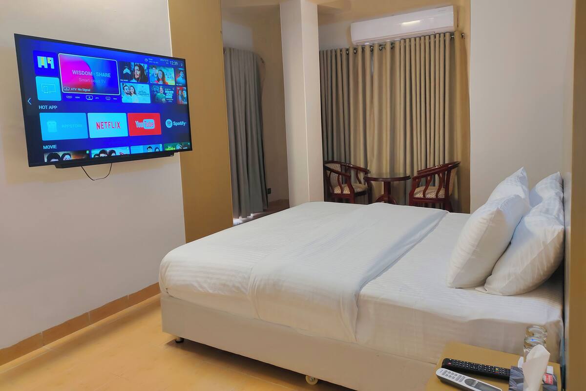 A hotel room with a bed, a TV mounted on the wall displaying various apps, a small table with a remote control, and a water bottle.