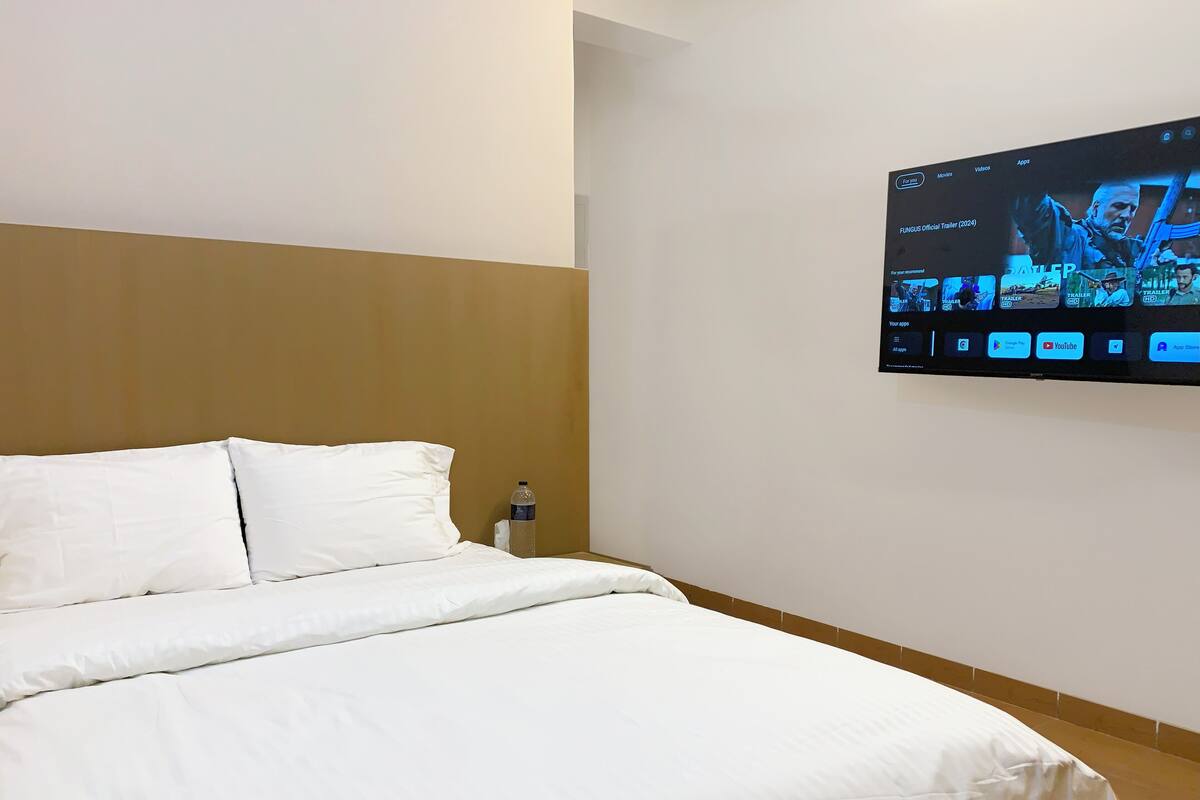 A hotel room with a bed, a water bottle on the nightstand, and a TV mounted on the wall displaying a movie.