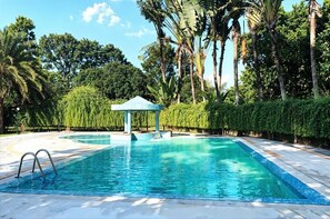 A rectangular swimming pool with a clear blue water surface, surrounded by lush greenery and palm trees. - Kazi Resort & Hotel (Kapasia)