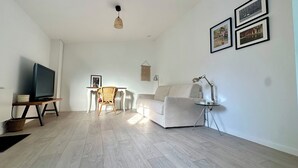 Un salon avec un téléviseur, un canapé blanc, un fauteuil en osier, une petite table et une lampe.