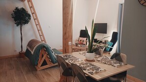 Ein gemütliches Wohnzimmer mit einem Holzesstisch, Stühlen, einer Leiter, einem Blumentopf und einem Fernseher an der Wand.