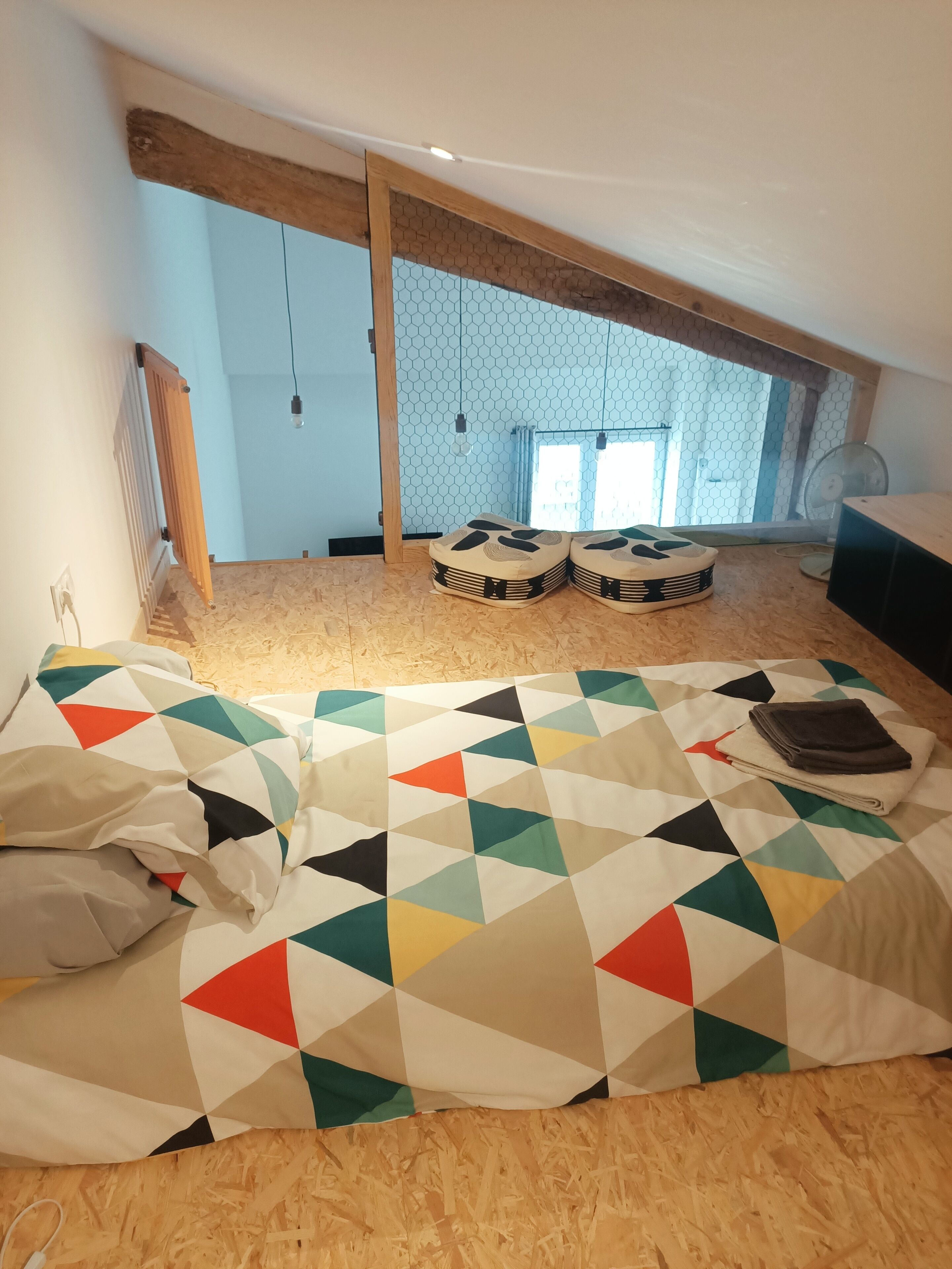 Una habitación con techo de madera inclinado, una cama con colcha de diseño geométrico y colores vivos, dos taburetes de madera y una pequeña mesa con una toalla doblada.
