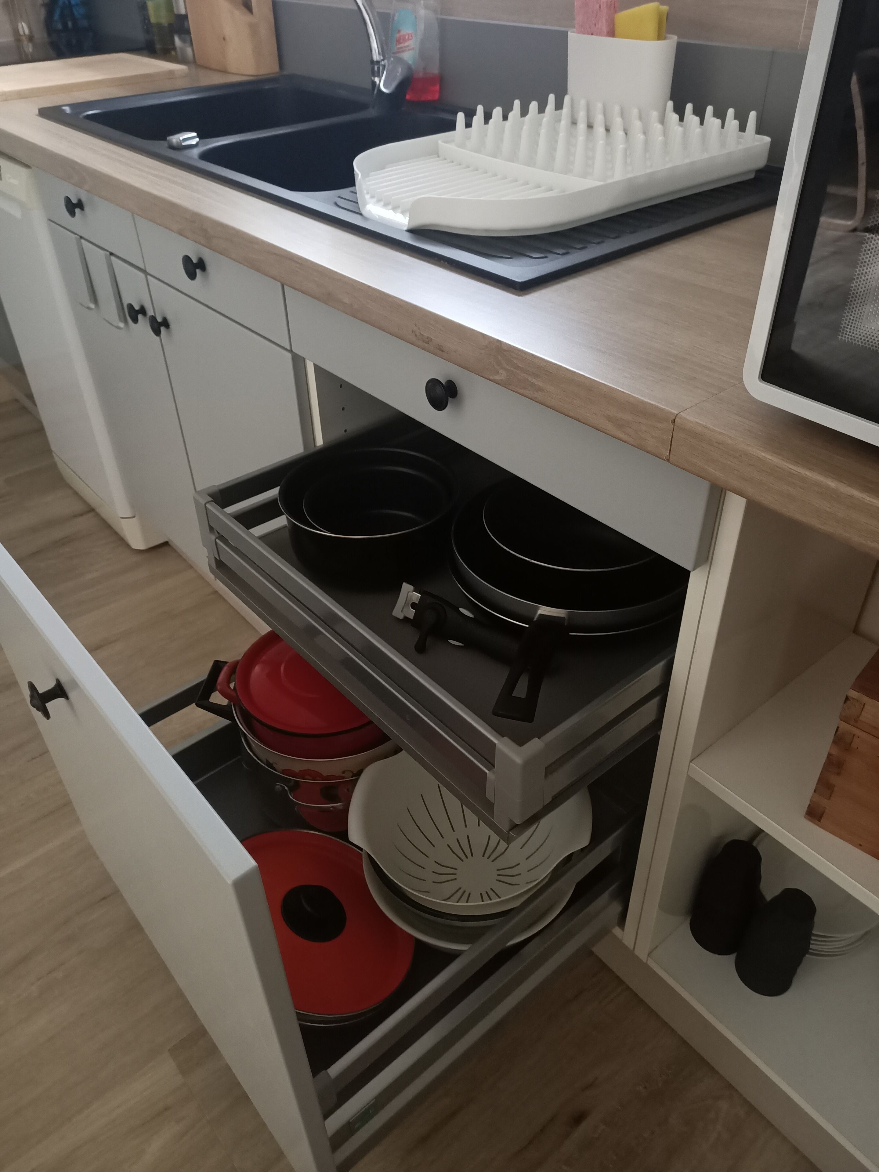 A kitchen with a sink, a white dish rack, and a drawer with pots and pans.