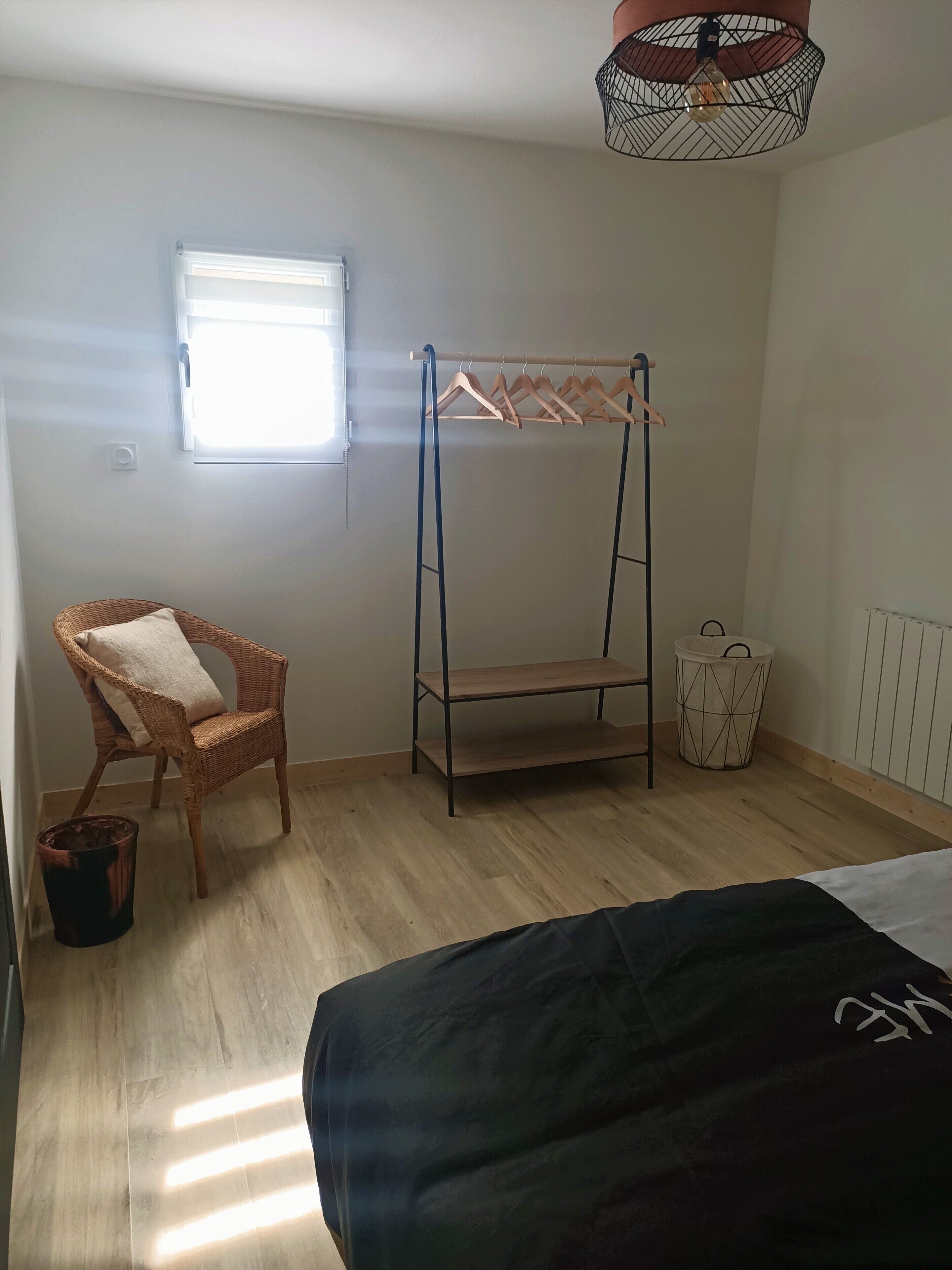Un dormitorio con una cama litera, una silla de mimbre, un ventanal pequeño, un radiador y una lámpara colgante.