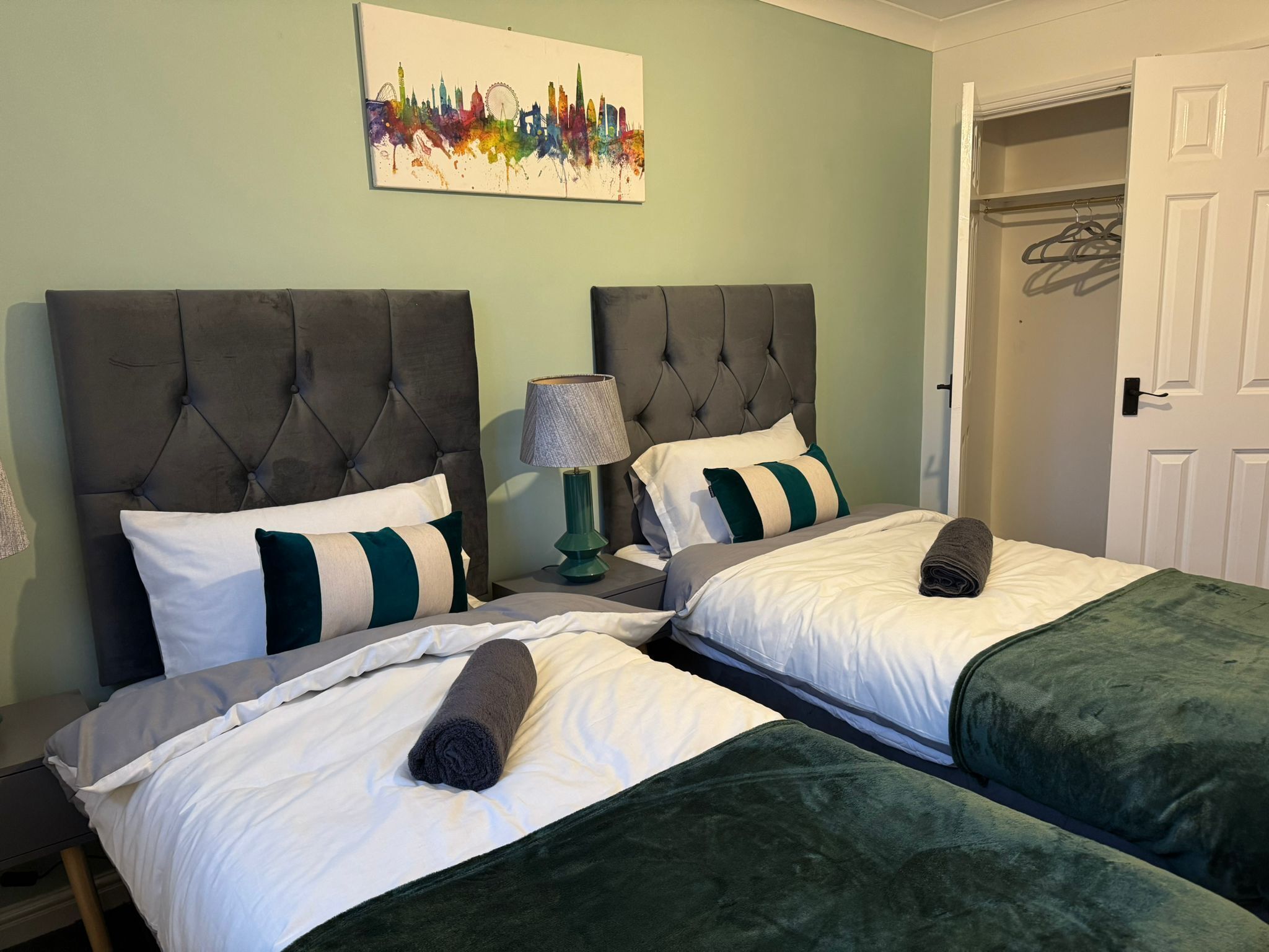 Une chambre d’hôtel avec deux lits, chacun doté d’une tête de lit grise, de draps blancs et de coussins décoratifs verts. Il y a une lampe sur un côté et une porte sur la droite.
