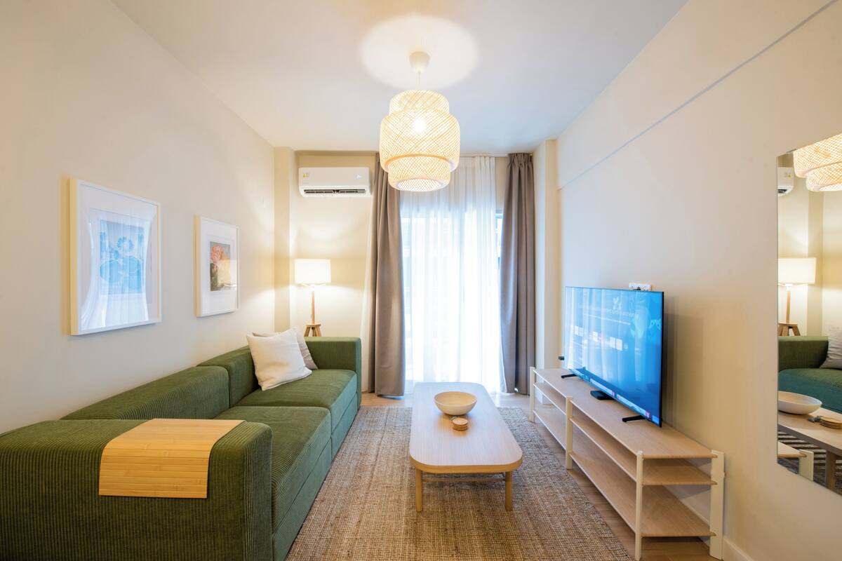 Ein modernes Wohnzimmer mit einem grünen Sofa, einem Holztisch, einem Flachbildfernseher und einem großen Fenster mit Vorhängen.