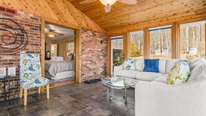 A cozy room with a wooden ceiling, a brick wall, and a large window. There's a bed, a sofa, and a glass coffee table. - Daisy Farm | Pool, Hot Tub & Animals 🐴 + Biltmore Pass Near Asheville (Candler)