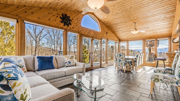 A spacious room with a wooden ceiling, large windows, a sofa, a glass coffee table, and a dining area with chairs. - Daisy Farm | Pool, Hot Tub & Animals 🐴 + Biltmore Pass Near Asheville (Candler)