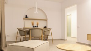 Una habitación moderna y minimalista con una mesa redonda de madera, dos sillones tapizados, un otoman beige y un estante en la pared con objetos decorativos.