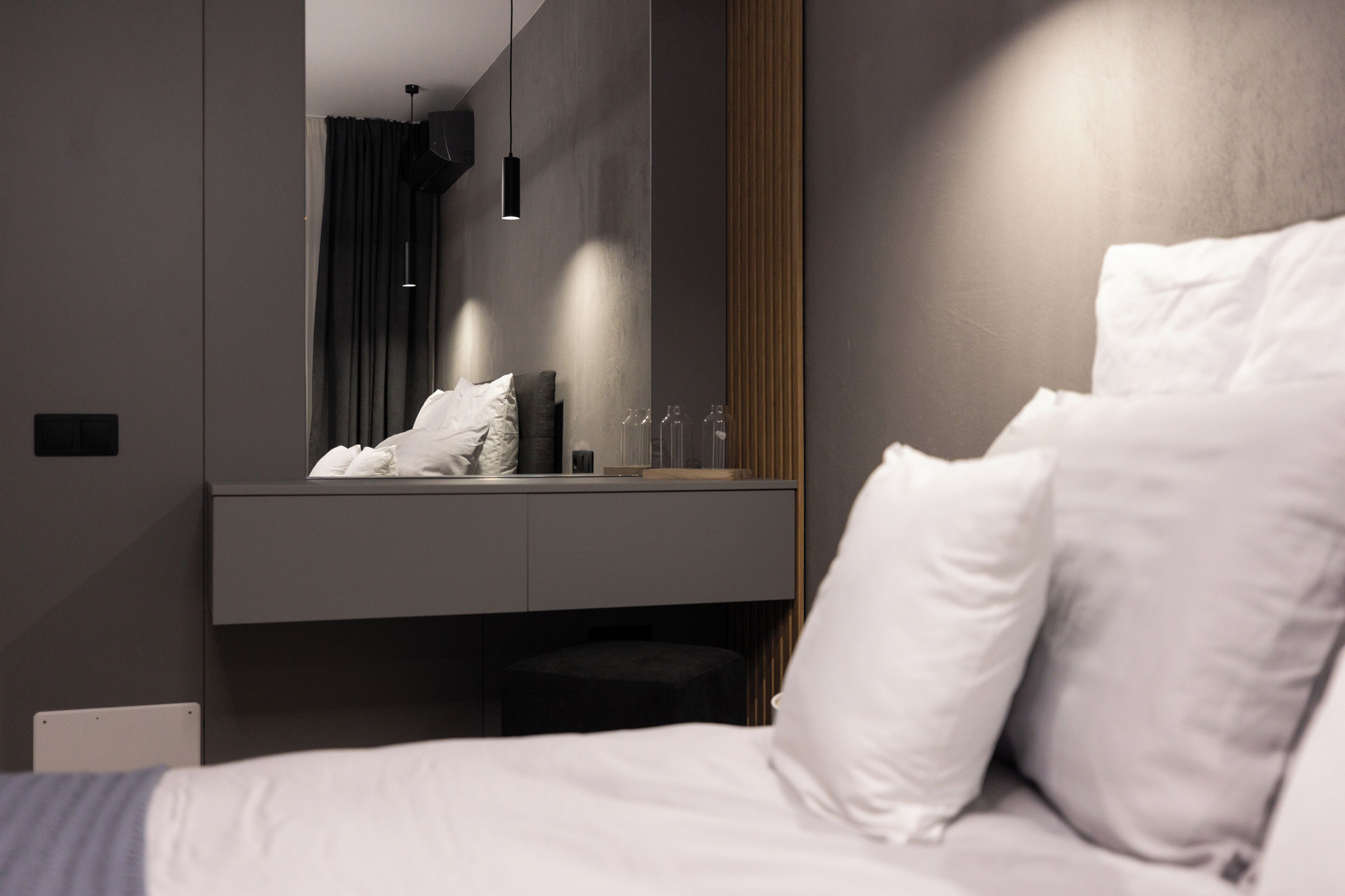 A modern hotel room with a large bed, a bedside table, a mirror, and a dark-colored wall.