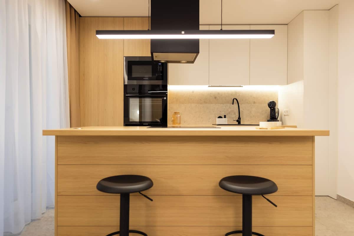 A modern kitchen with wooden cabinetry, a built-in oven, a microwave, and two black bar stools.