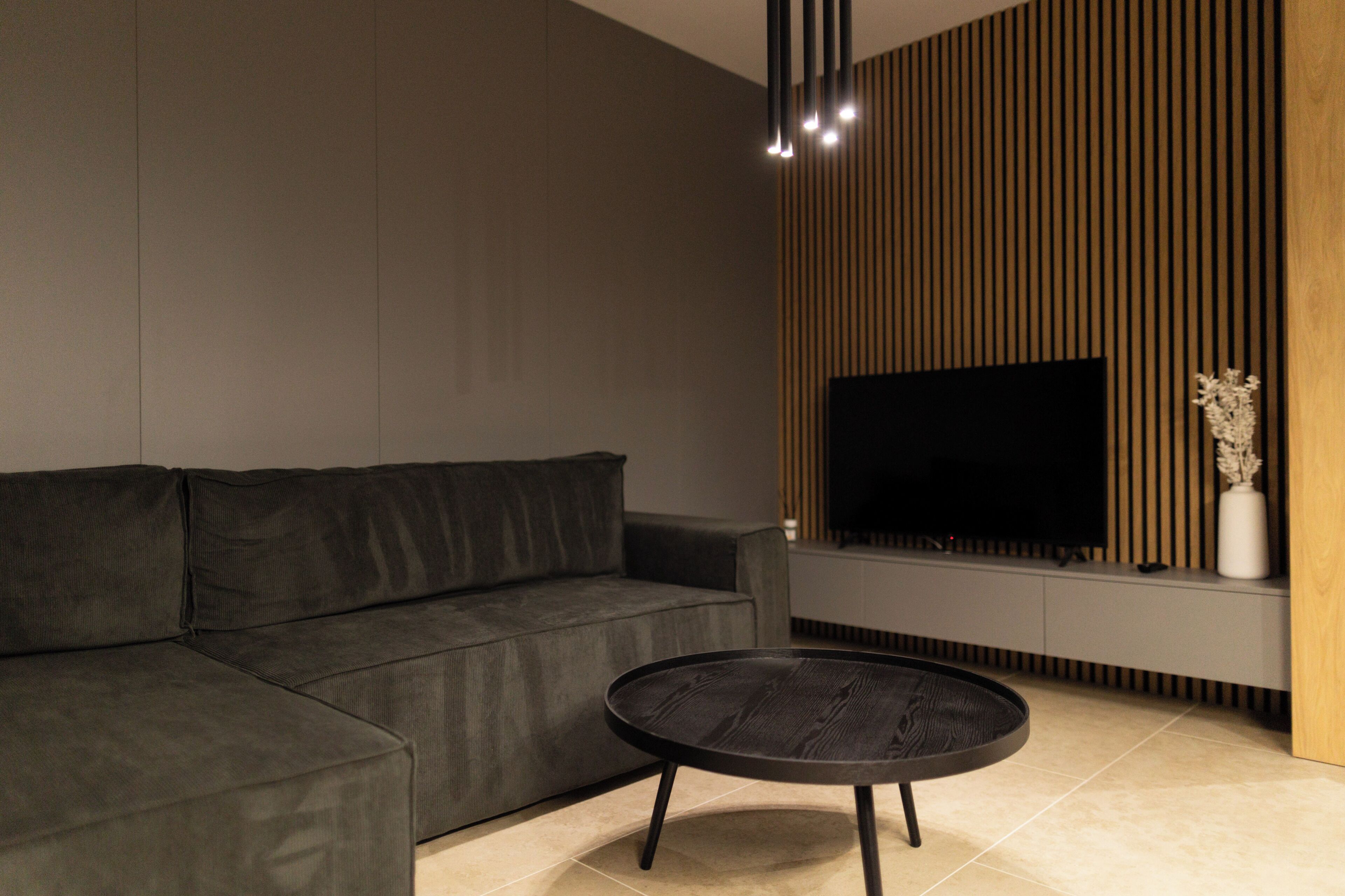 A modern living room with a sectional sofa, a round coffee table, a flat-screen TV mounted on a wooden wall, and a minimalist decor style.