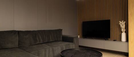 A modern living room with a sectional sofa, a round coffee table, a flat-screen TV mounted on a wooden wall, and a minimalist decor style.