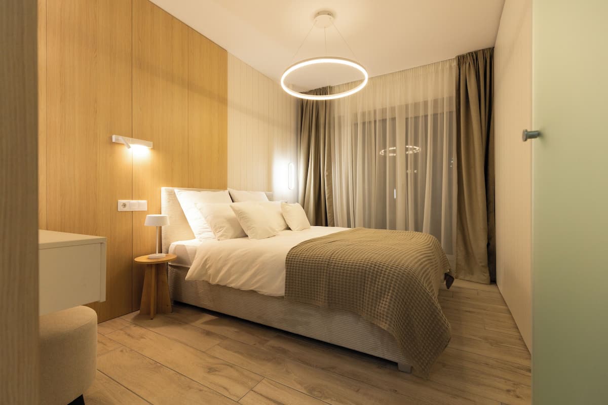 A modern bedroom with a wooden floor, a bed with a beige blanket, a bedside table with a lamp, and a large window with sheer curtains.