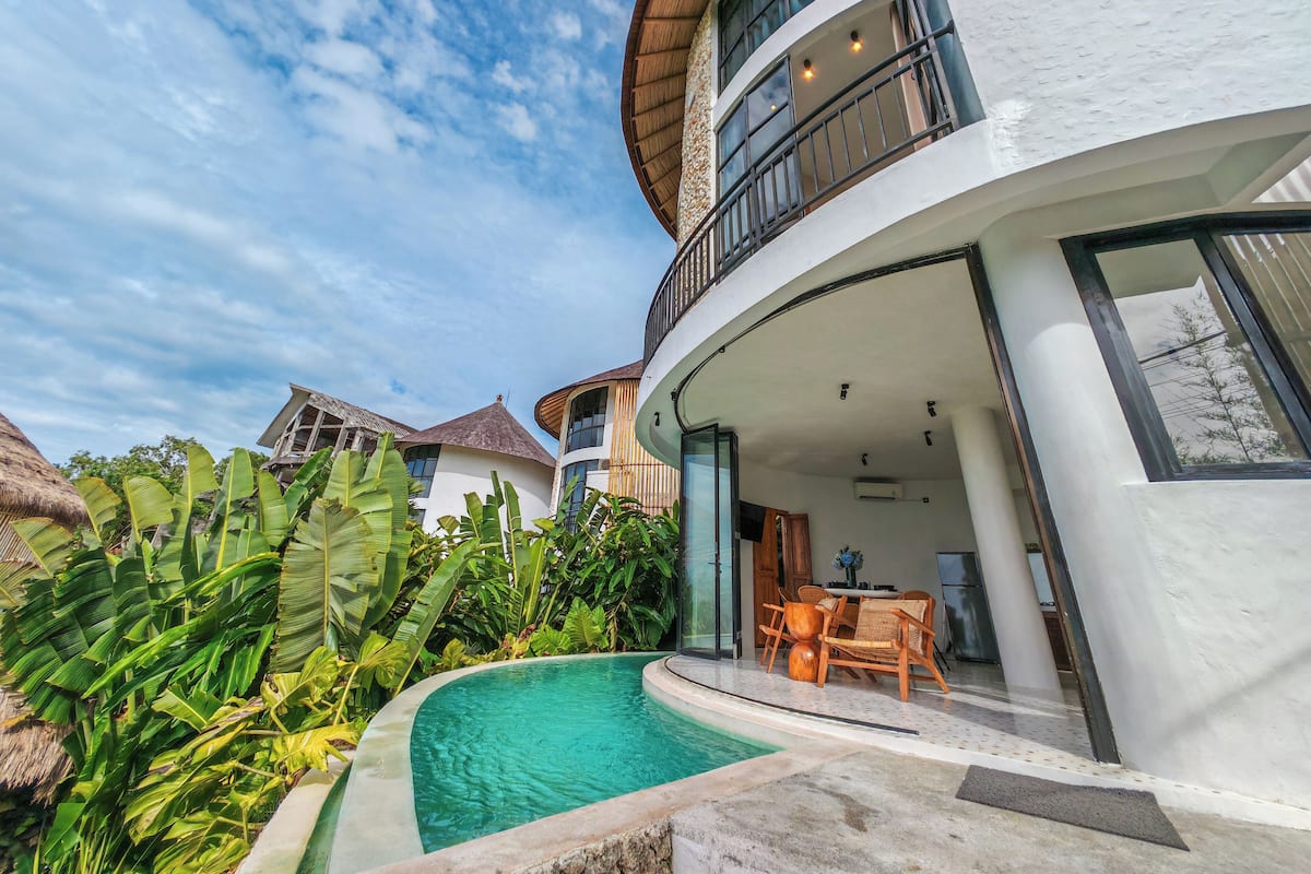 A modern resort with a pool, outdoor seating, and a thatched-roof structure.