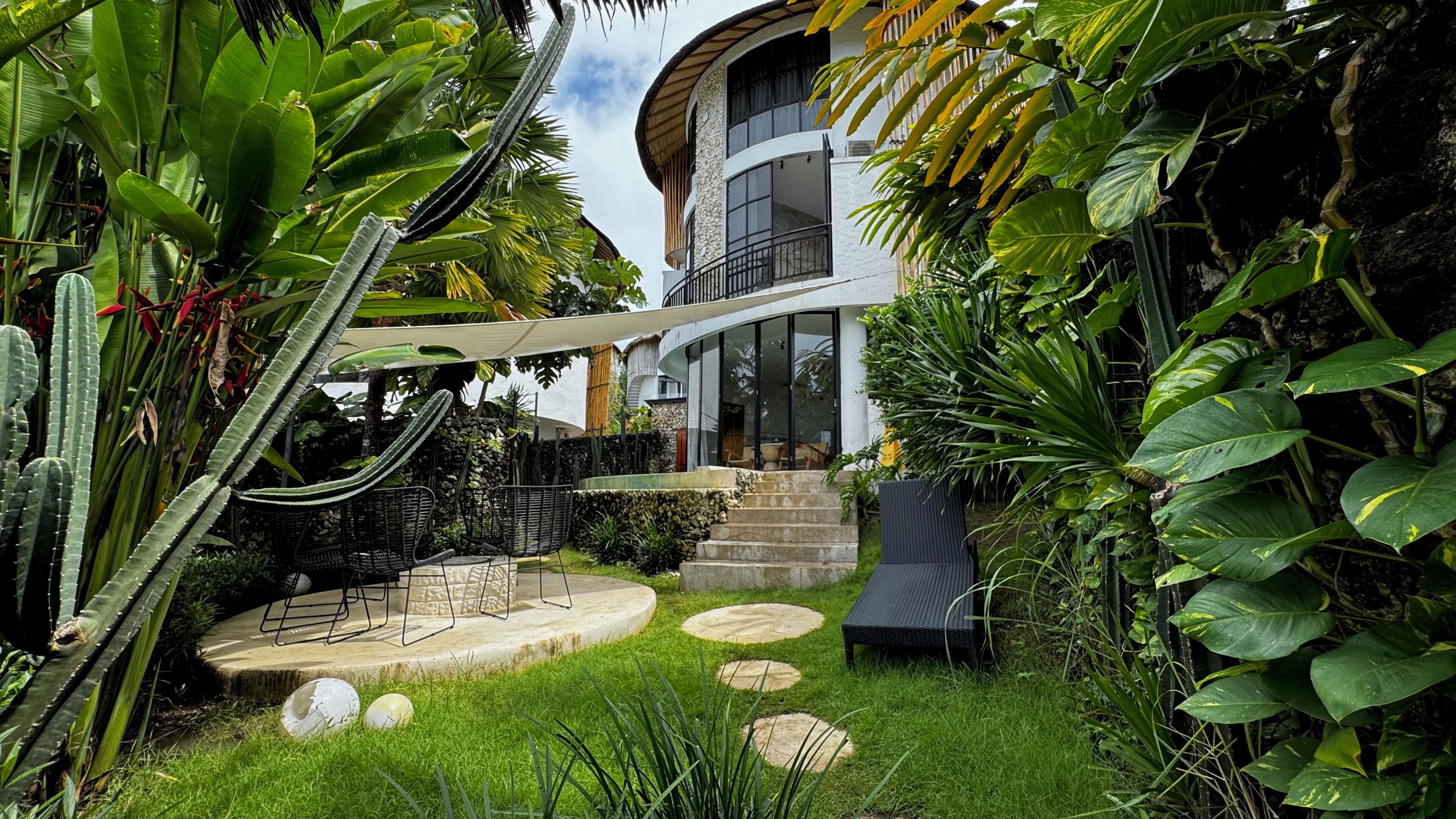 A modern house with a curved balcony, surrounded by lush tropical plants and a stone pathway leading to the entrance.