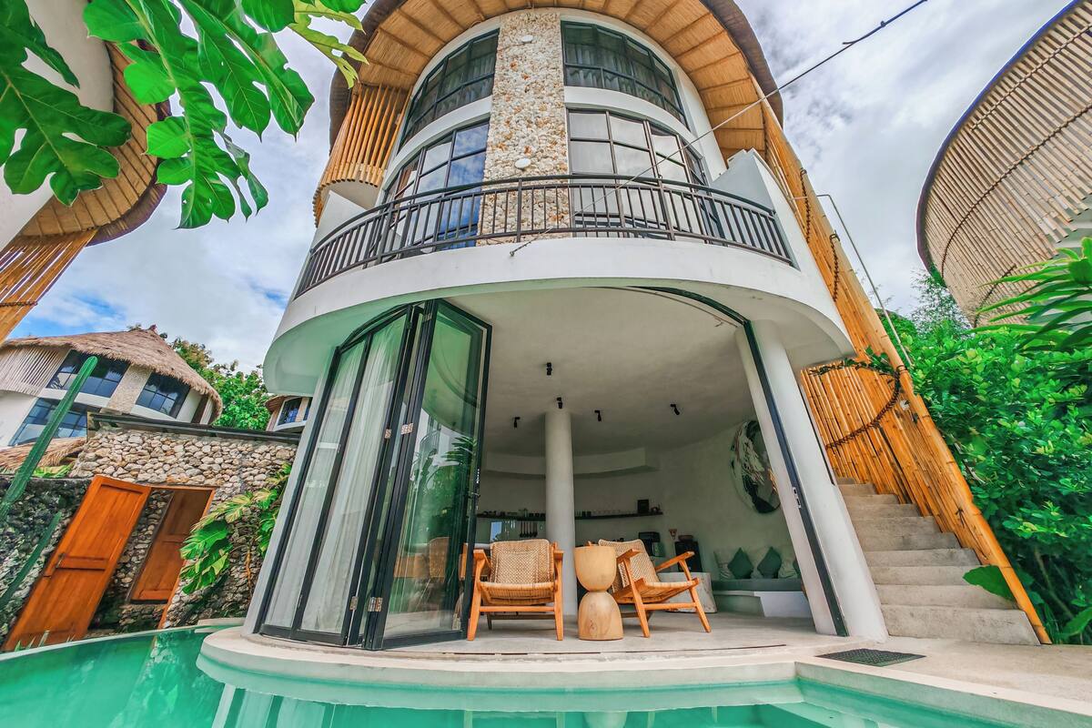 A modern two-story house with a curved balcony, a pool, and a stone wall.