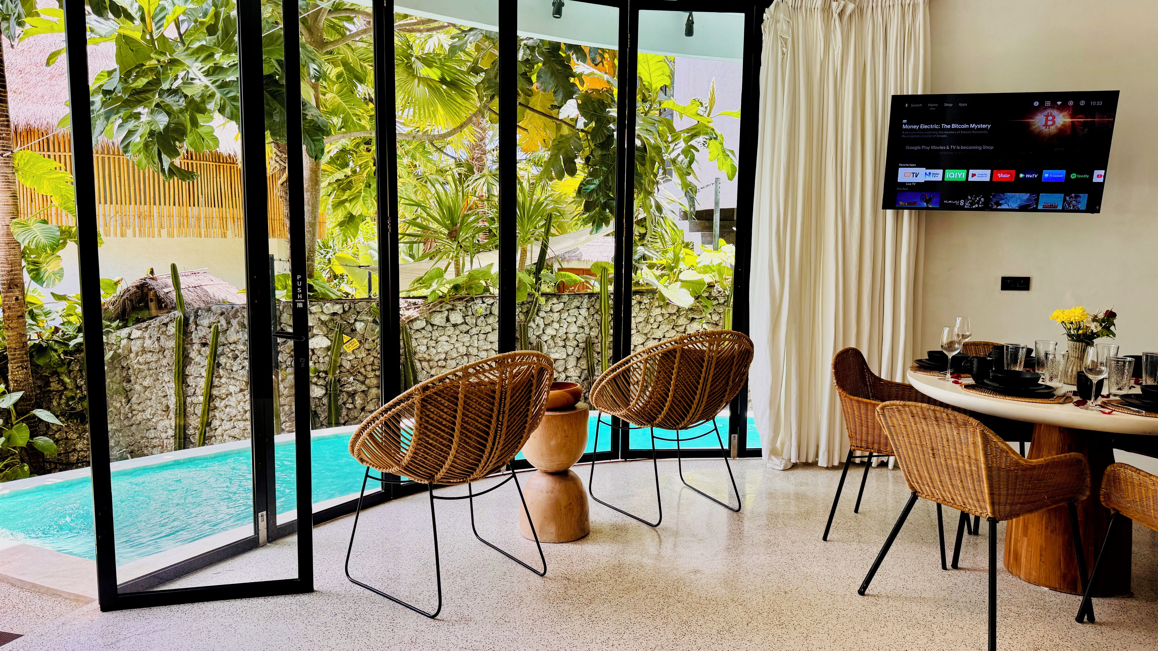 A modern room with a pool, wicker chairs, a dining table set for four, and a TV displaying a program.