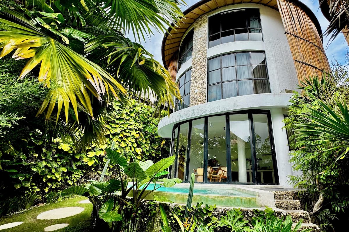 A modern building with a curved facade, large windows, and a pool area surrounded by lush tropical vegetation.
