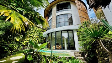 A modern building with a curved facade, large windows, and a pool area surrounded by lush tropical vegetation.