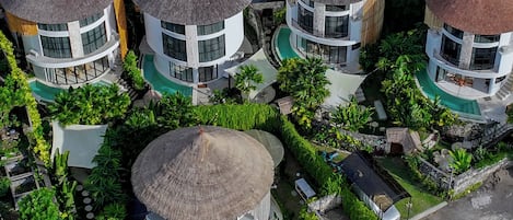 A cluster of circular buildings with thatched roofs, surrounded by lush greenery and a swimming pool.
