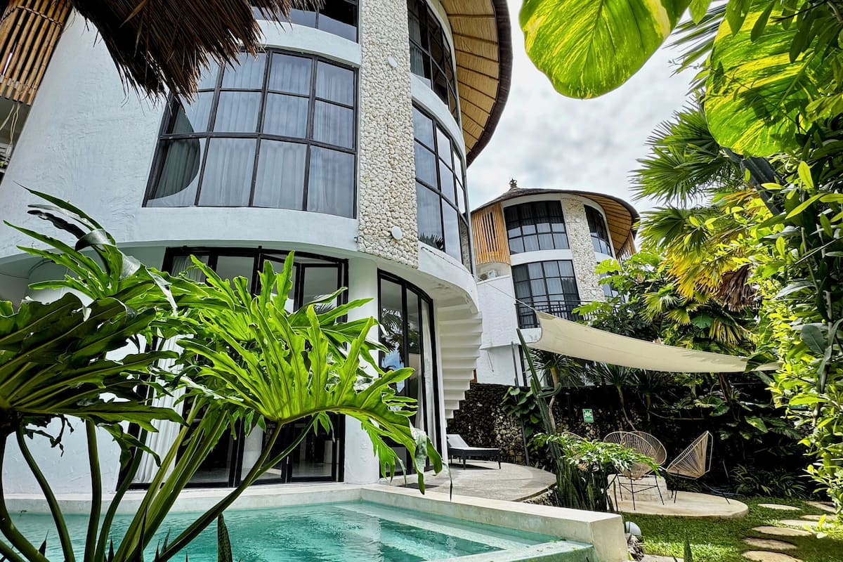 A modern two-story building with a pool, surrounded by lush greenery and tropical plants.