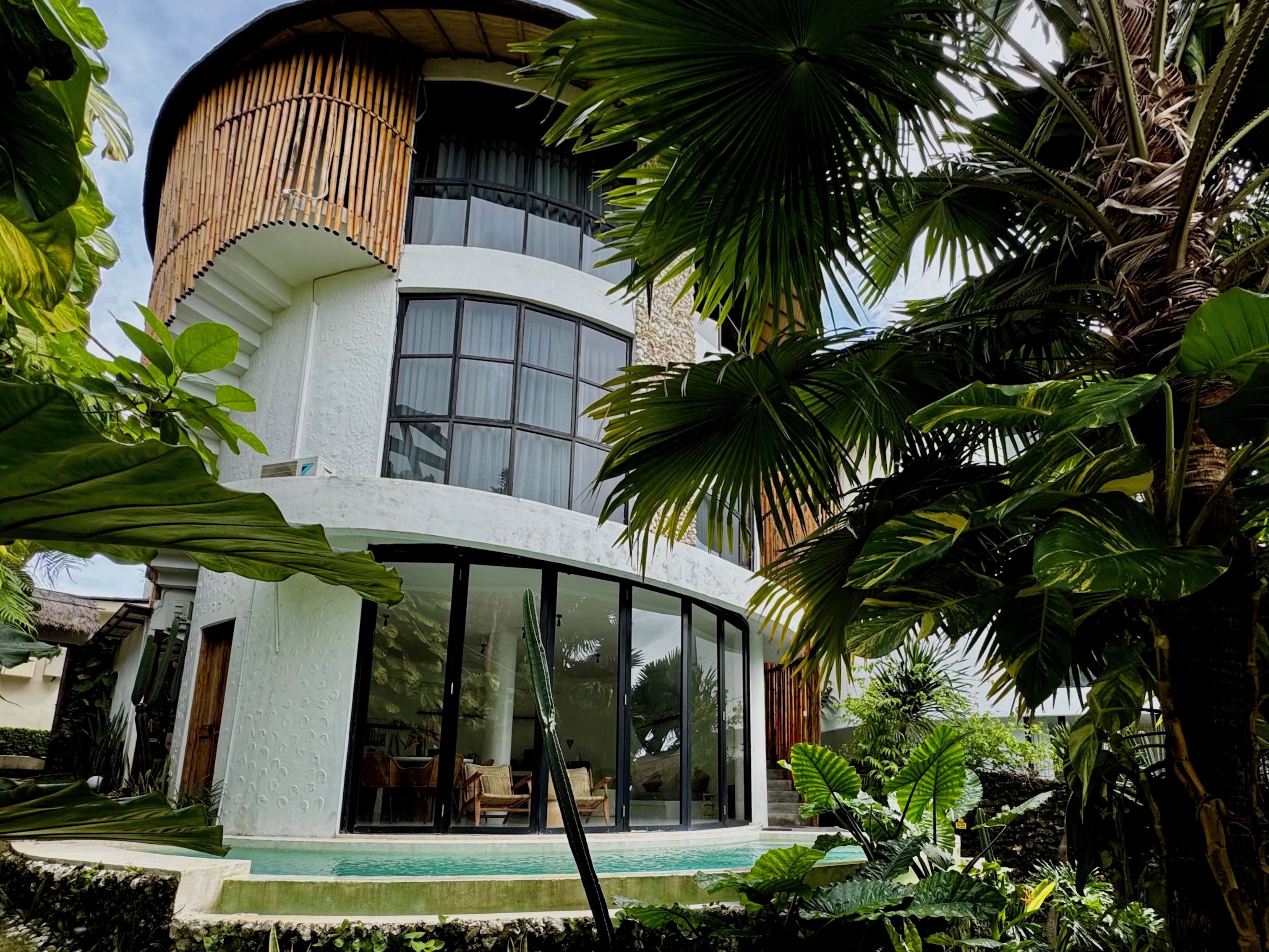 A modern house with a curved design, large windows, and a pool area surrounded by lush greenery.